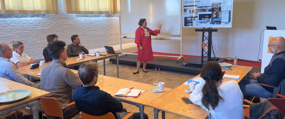 Presenter in red dress points to whiteboard during a professional meeting with attentive audience.