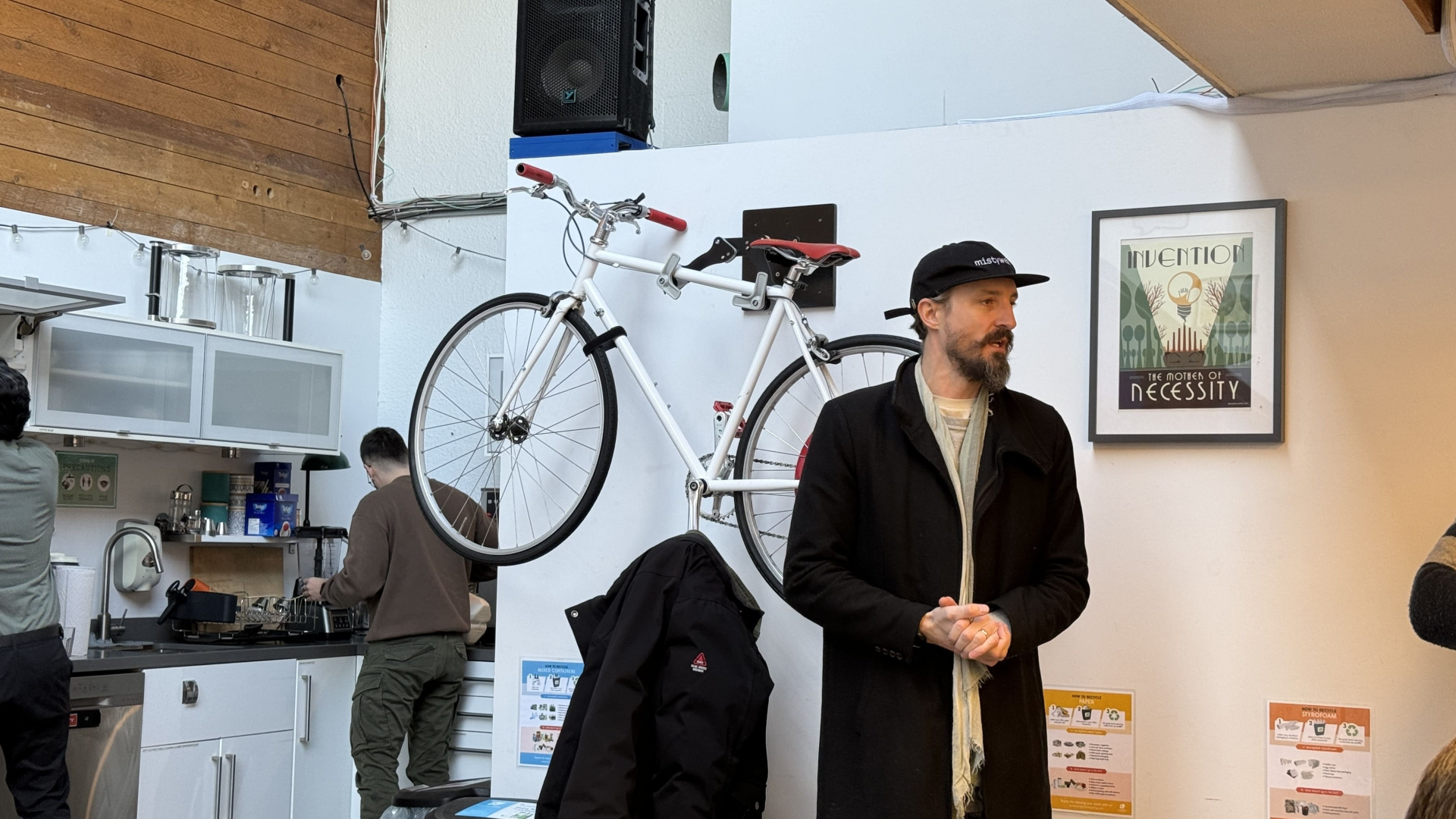 Man in black coat and cap speaking, white bicycle mounted on wall.