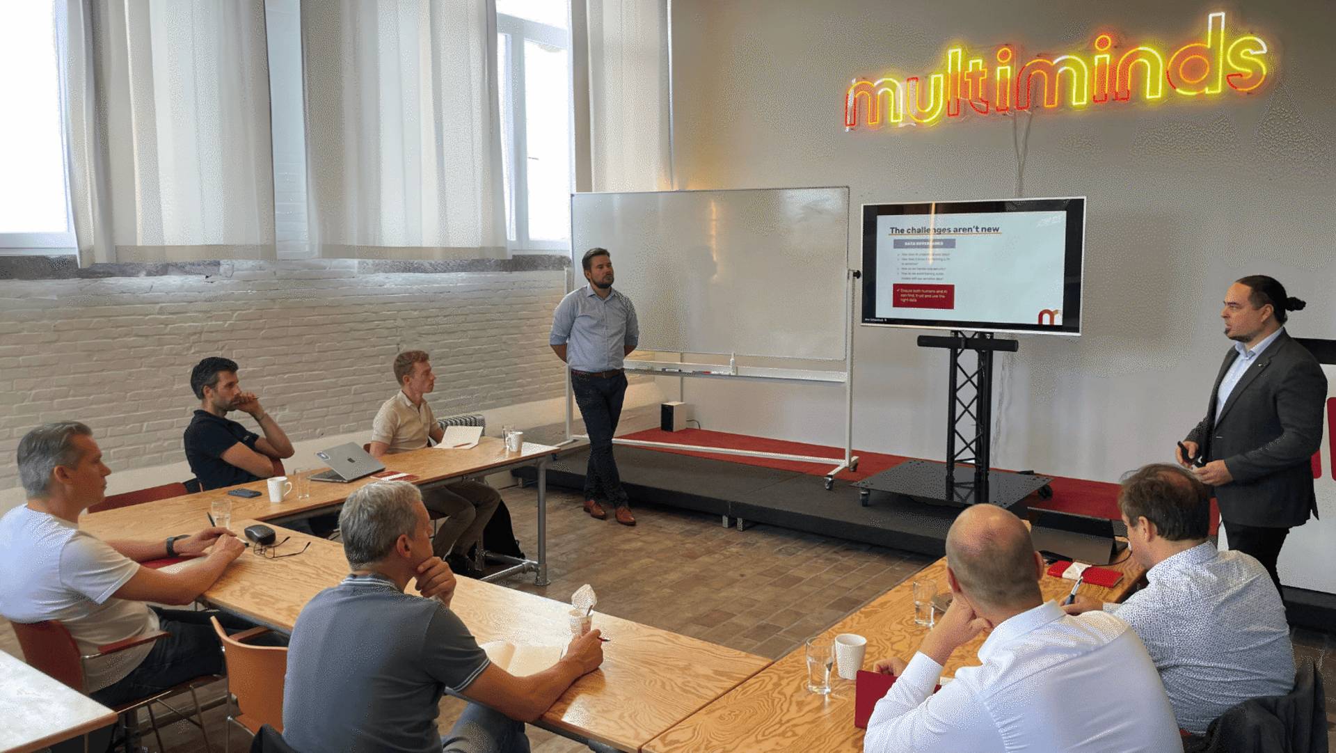 Attendees listening to a "Multiminds" presentation with one speaker near a screen displaying "The challenges aren't new."