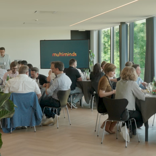 Attendees at a business event, seated at tables with a "multiminds" logo on a screen in the background.