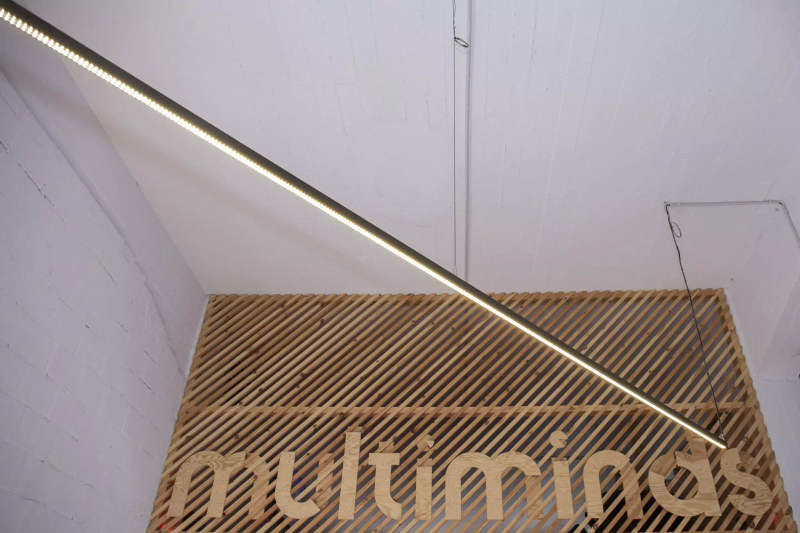 Linear LED light fixture illuminating a wooden lattice wall with the "multiminds" logo.