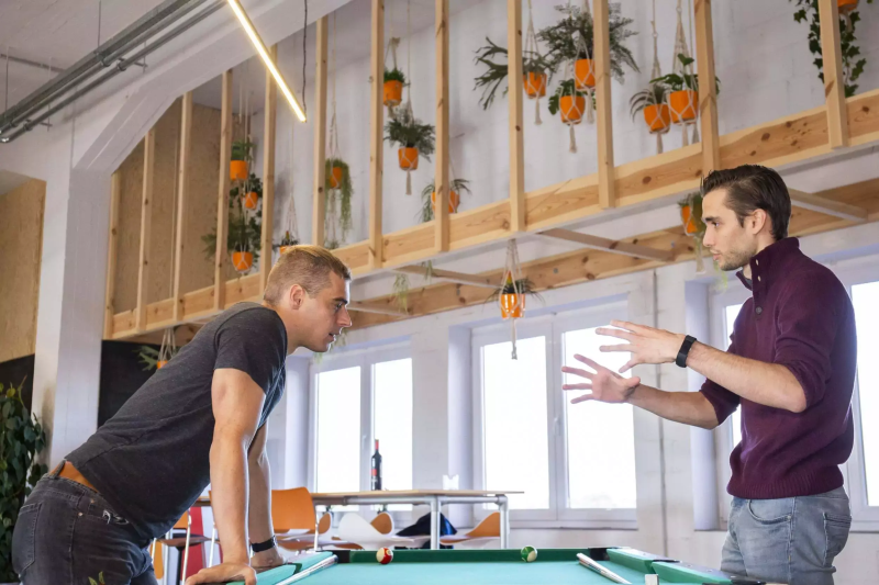 Nicolas and Geoff discussing strategy around a pool table, modern office space with hanging plants in the background.